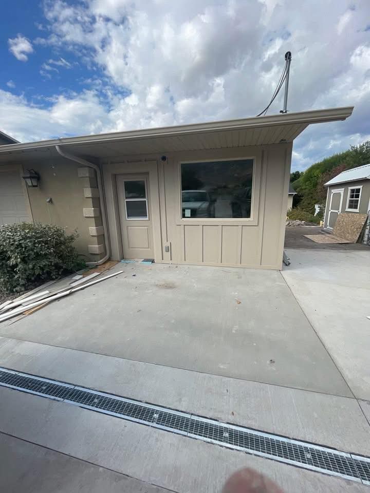 Tan building exterior with a door and window, a concrete patio, and a drainage grate in the foreground.