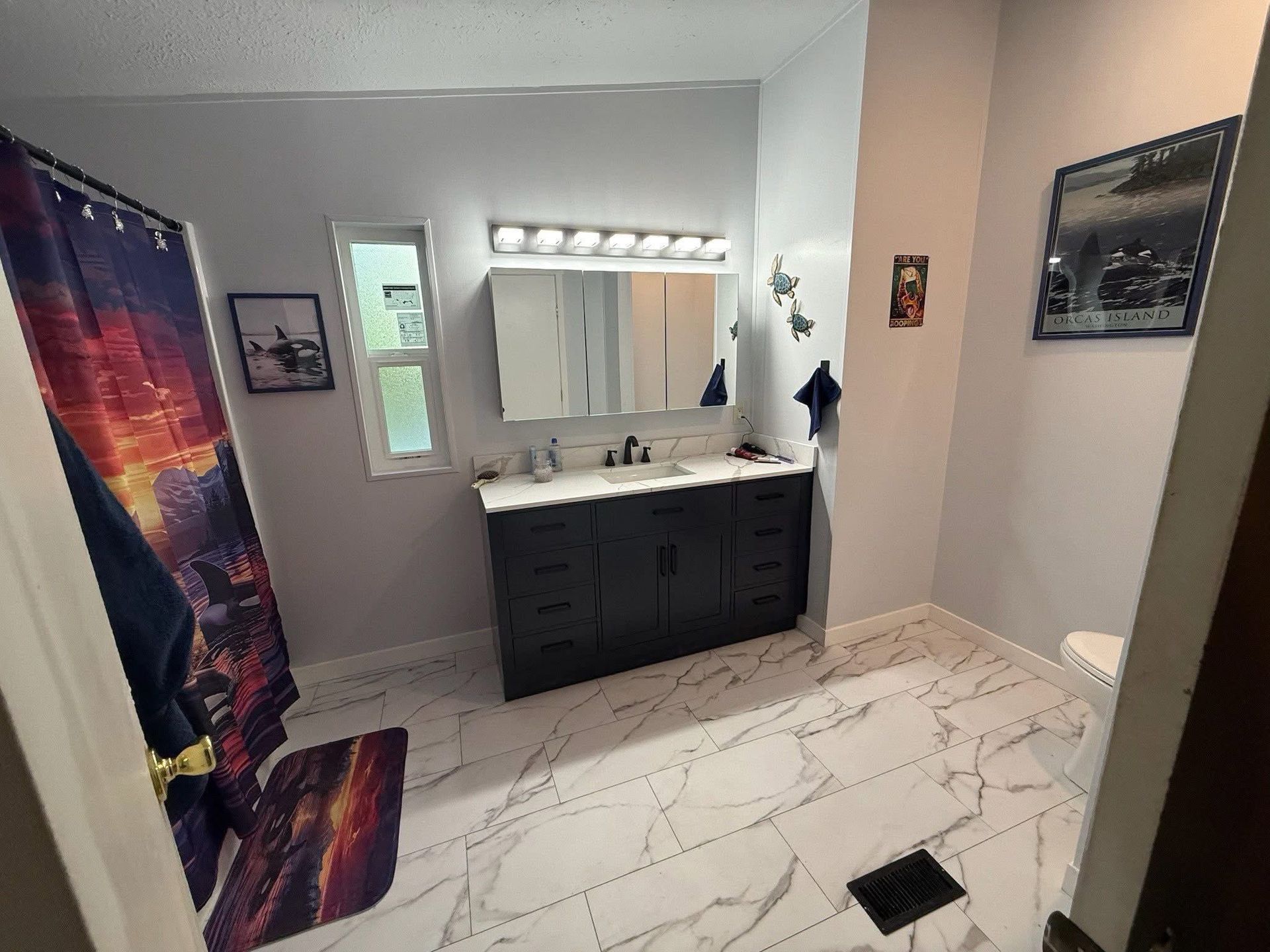 A modern bathroom featuring a dark vanity, marble-patterned tile floor, a shower with a colorful curtain, and wall art.