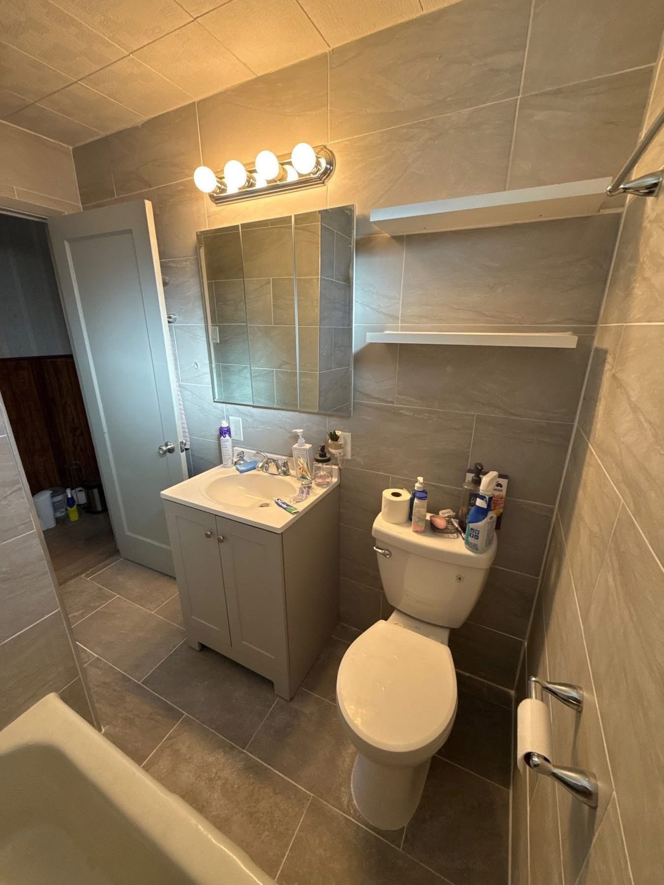 Small bathroom with grey tiled walls and floor, a white vanity, a toilet, a square mirror, and two open wall shelves.