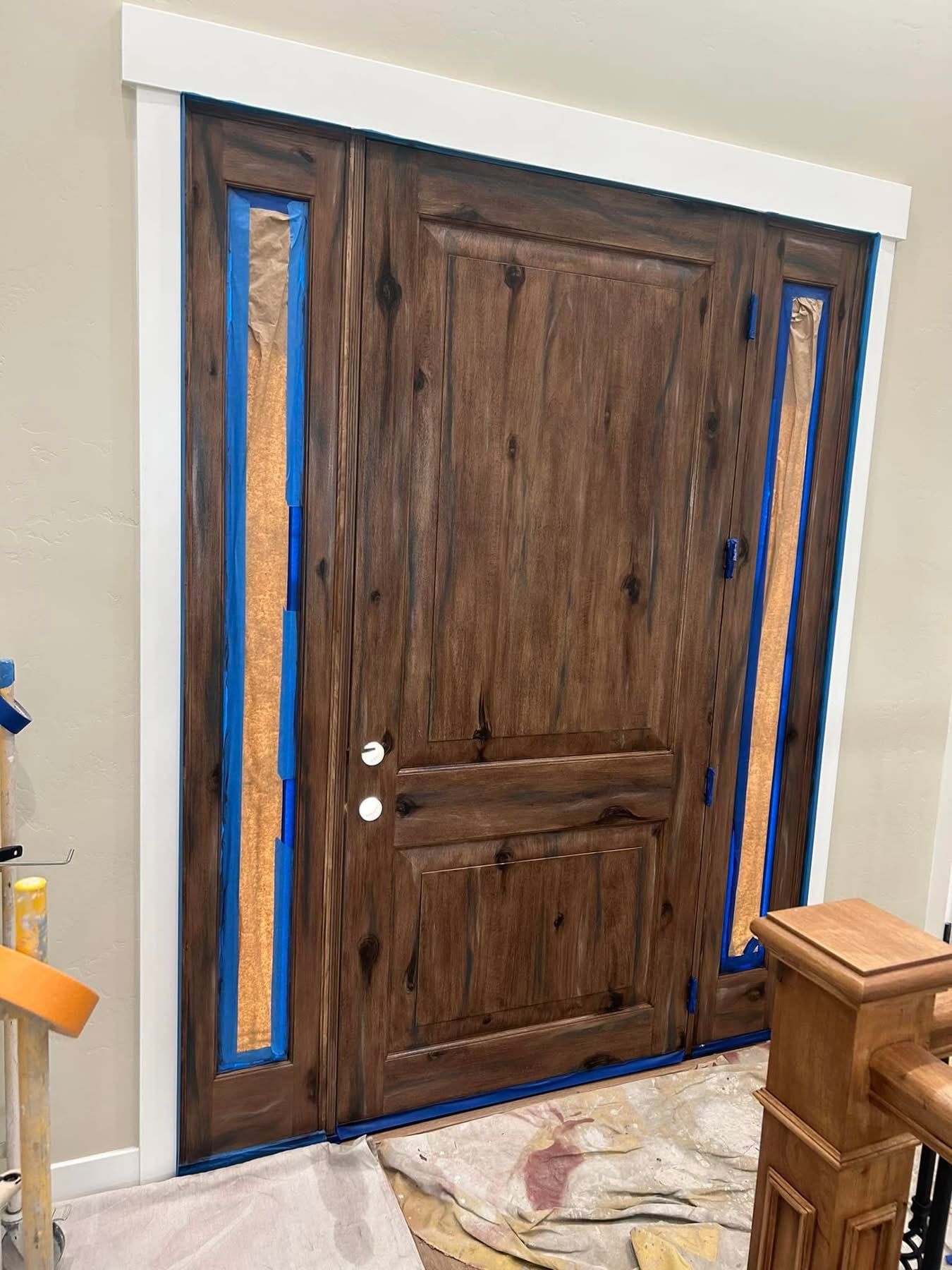 A dark wood front door with sidelights is taped off for painting, surrounded by a white frame in an indoor home setting.