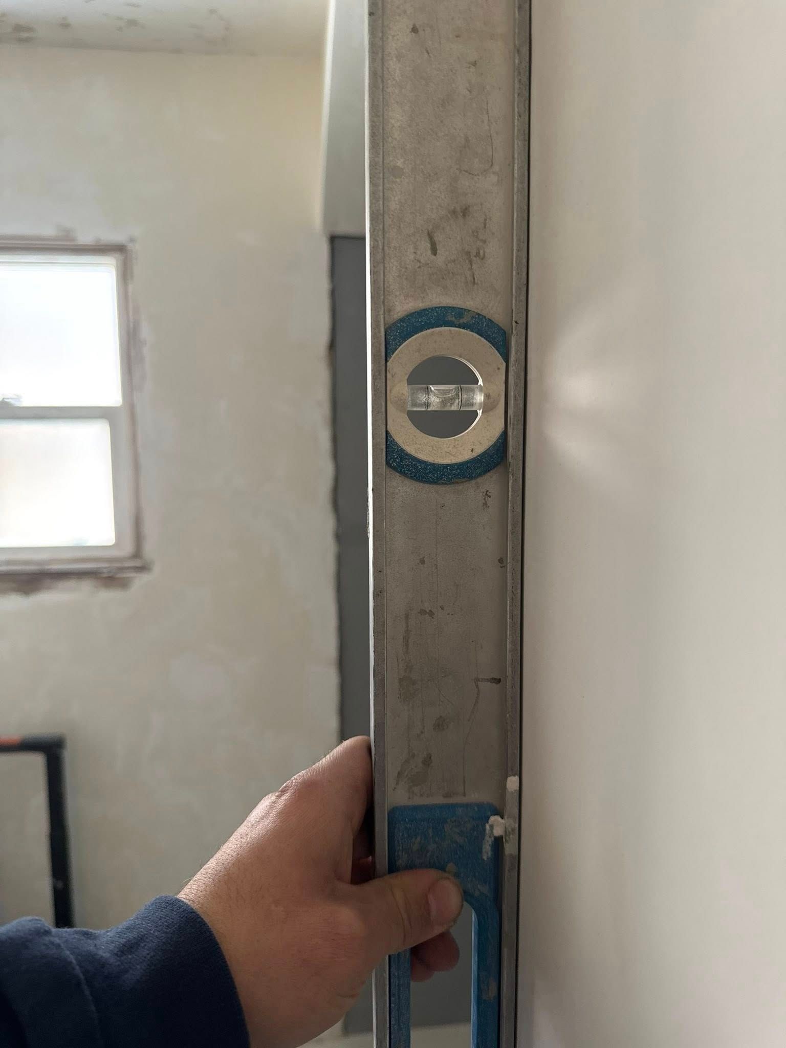 A person holds a metal level against a wall to check for vertical plumbness. The level's bubble shows the wall is not level.