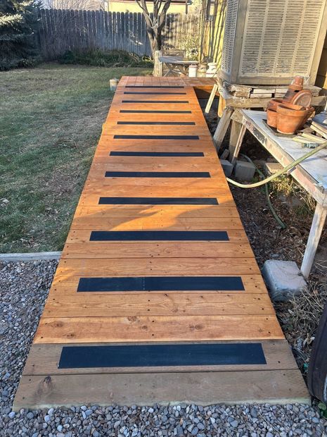 A wooden ramp with horizontal black non-slip treads leading from gravel toward a wooden platform in a yard.