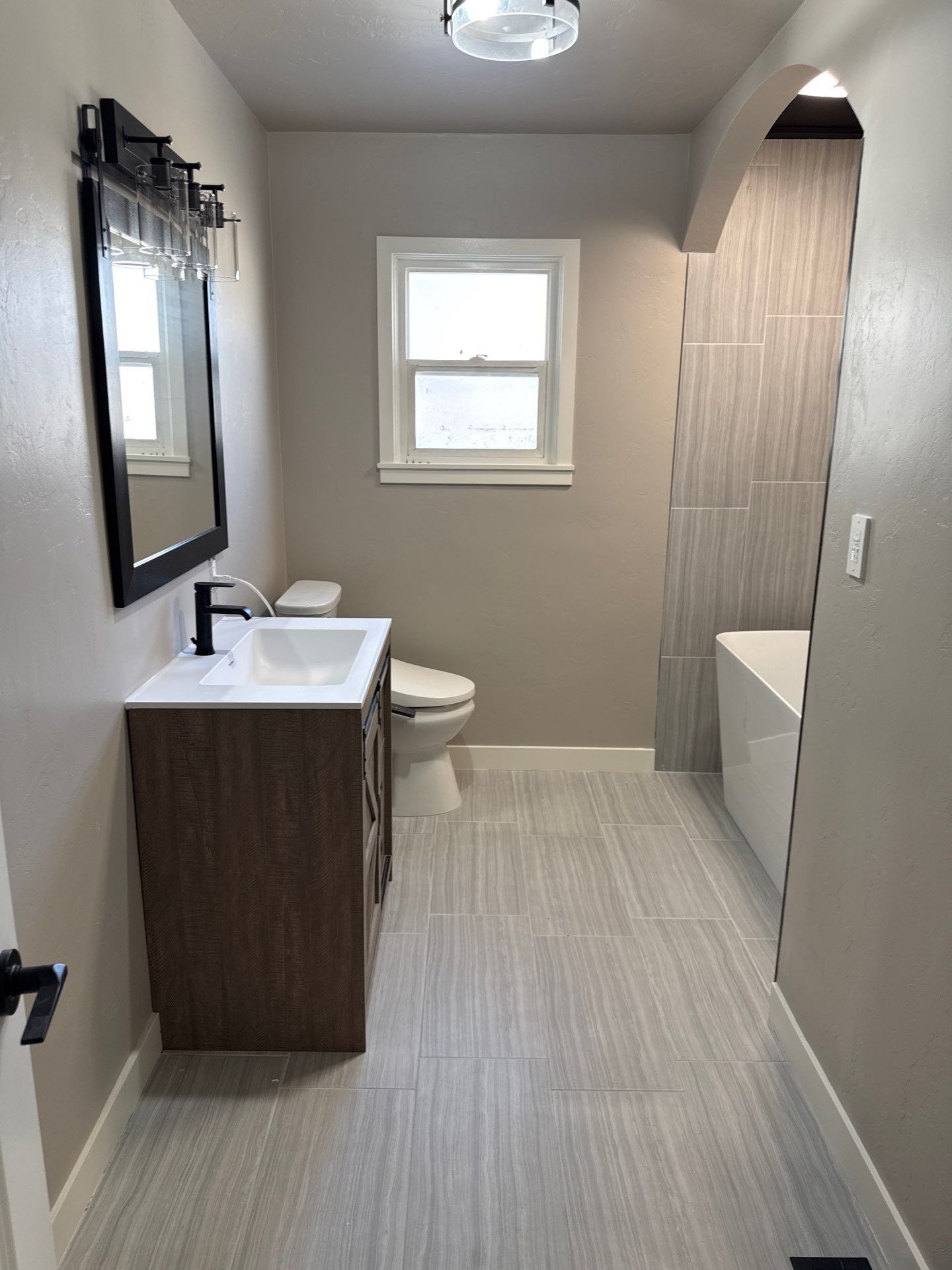 A bathroom with a wood-toned vanity, white sink, black mirror, toilet, and tiled floor leading to a bathtub alcove.