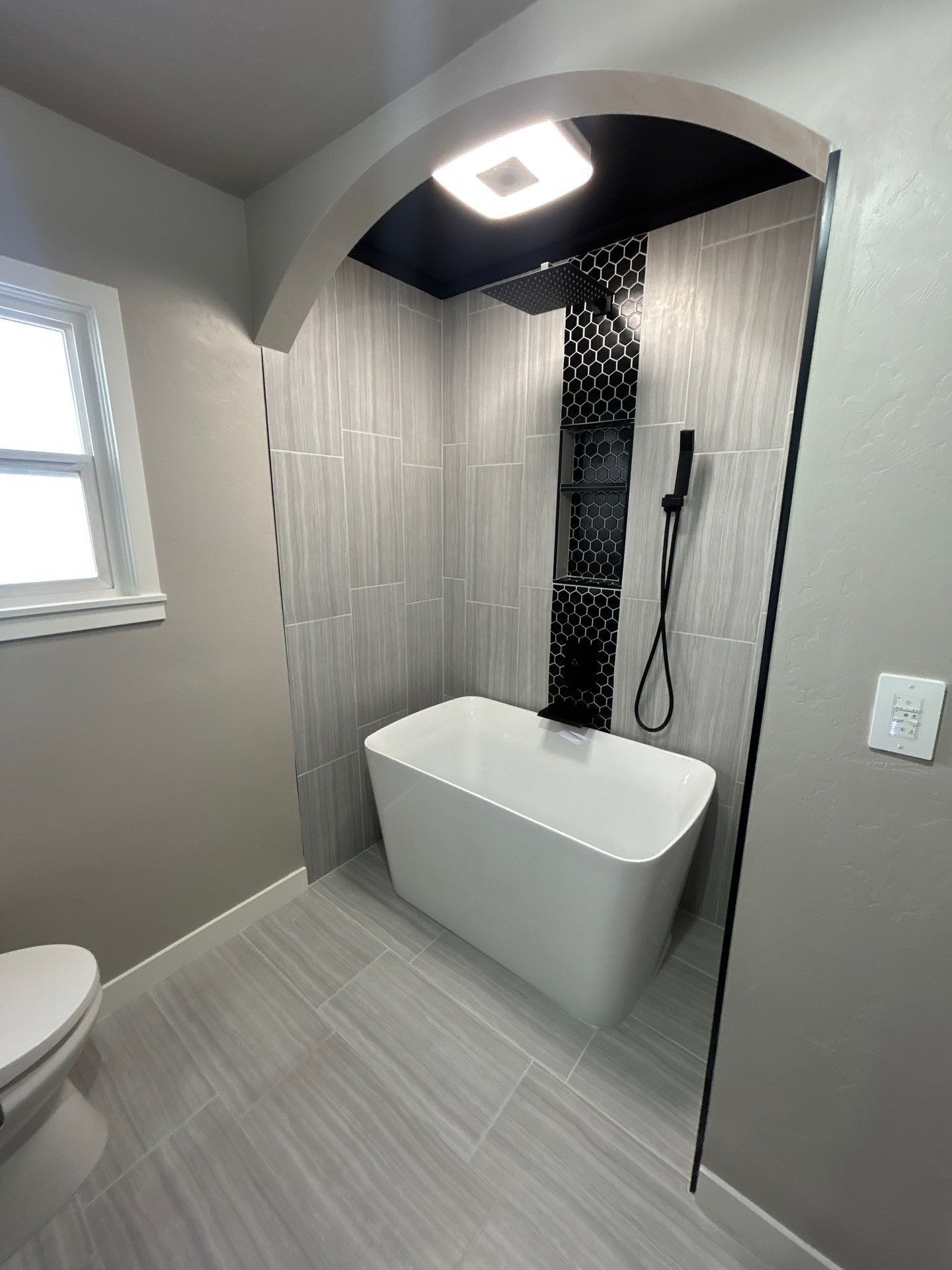 A freestanding white bathtub sits beneath an arched alcove with grey tiled walls and a black mosaic vertical accent strip.