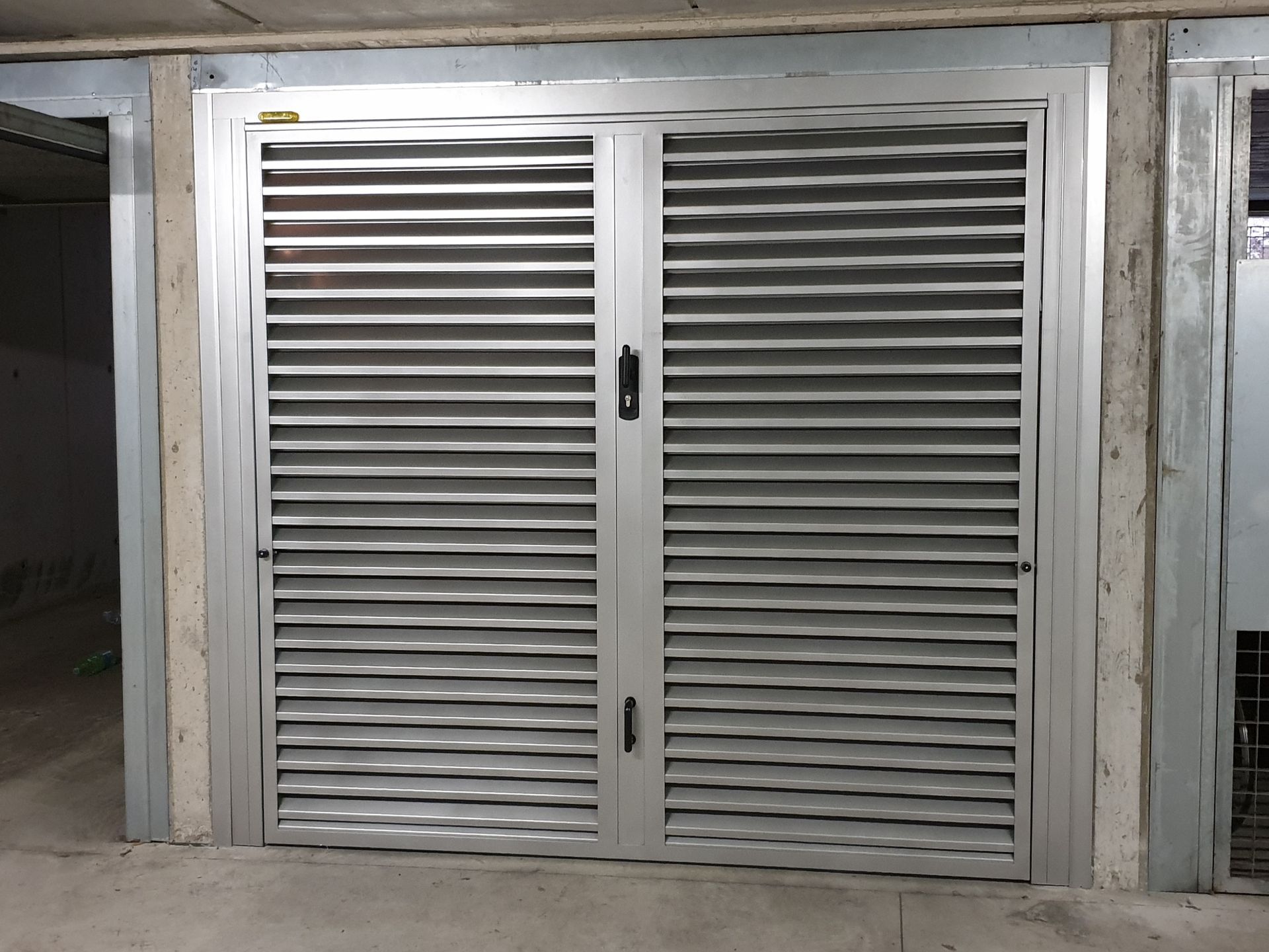 Porte a doppia persiana in metallo grigio incastonate in una parete di cemento, come quelle di un garage o di un ingresso di servizio.