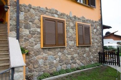 Esterno di una casa in pietra con la parte superiore della parete arancione, due persiane in legno e un piccolo cortile recintato.