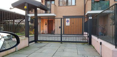 Ingresso del condominio con cancello, recinzione metallica, cortile pavimentato e porta con cartello al centro.