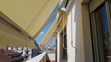 Balcone con tende da sole beige, palazzi e cielo azzurro accanto a una parete esterna bianca.