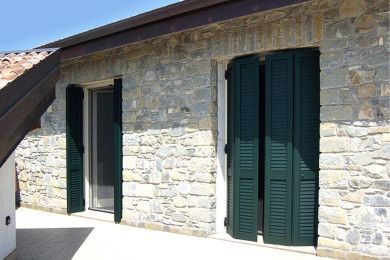 Muro di pietra della casa con persiane verde scuro e un ingresso illuminato dal sole.