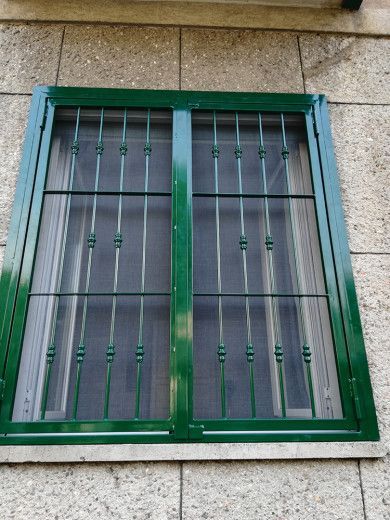 Griglia di sicurezza in metallo verde su un muro di pietra, con tende chiuse alle sue spalle.