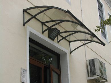 Tenda da sole metallica curva sopra un ingresso di un edificio di colore chiaro, con un condizionatore d'aria montato accanto.