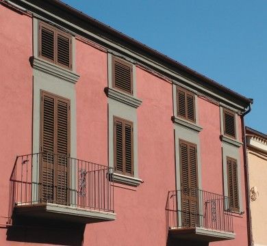 Edificio in stucco rosa con finestre con persiane e balconi in ferro battuto sotto un cielo azzurro.