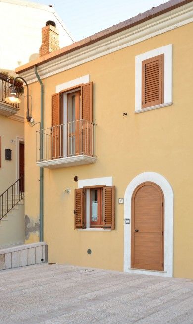 Facciata gialla della casa con finiture bianche, persiane in legno, balcone e porta d'ingresso ad arco.
