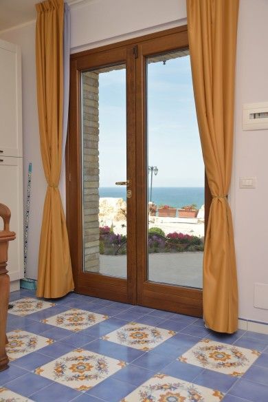 Porte finestre con tende color senape che si aprono su una vista sul mare, sopra un pavimento di piastrelle decorate blu e bianche.
