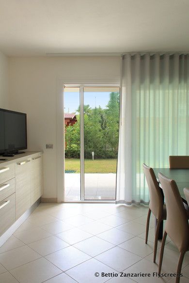 Luminoso soggiorno moderno con porte scorrevoli in vetro, tende bianche, TV, cassettiera e sedie da pranzo.