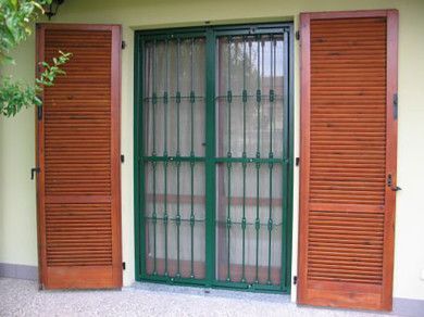 Porta verde con sbarre di sicurezza in metallo, affiancata da due persiane aperte in legno marrone su una parete verde chiaro.