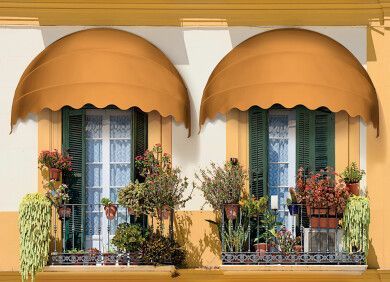 Due finestre del balcone con tende da sole arancioni e piante in vaso sulla facciata gialla dell'edificio.