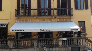 Terrazza del caffè con tenda da sole bianca, loghi di birra e balcone decorato su un edificio giallo.