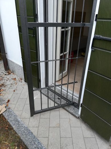 Cancello metallico aperto in uno stretto ingresso esterno con patio piastrellato e pannelli a parete verdi