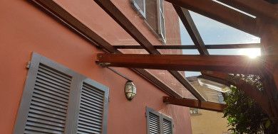 Edificio in stucco rosso con persiane grigie e pergolato in vetro sovrastante, sullo sfondo di un cielo azzurro.