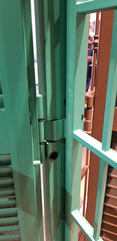 Cancello con chiavistello in metallo verde e recinzione, con uno scorcio di scale in legno sullo sfondo.
