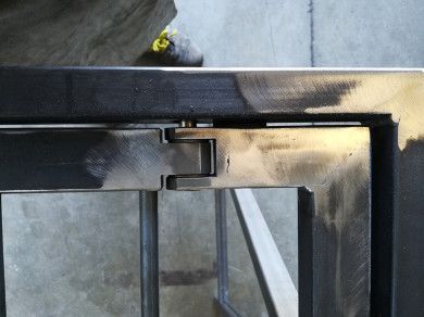Primo piano di una cerniera di una cornice in metallo nero, illuminata dalla luce del sole, con un piccolo fiore giallo sullo sfondo.