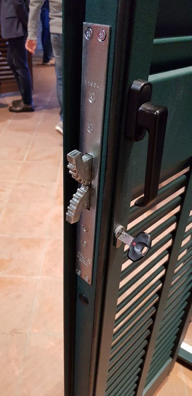 Primo piano di un cancello di metallo verde con chiavistello e lucchetto argentati, parzialmente aperto su un pavimento piastrellato.