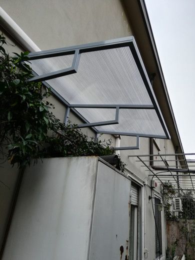 Passaggio pedonale esterno coperto con tettoia traslucida con struttura metallica, adiacente a un edificio bianco e circondato da vegetazione.