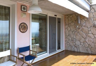Patio con porta scorrevole in vetro, parete rosa, sedia blu e muretto in pietra con vista sul mare.