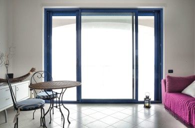 Luminoso soggiorno con porte scorrevoli in vetro blu, tavolino e sedie, e un divano viola sulla destra.