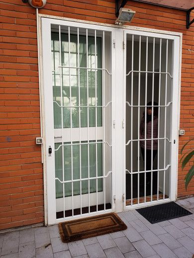 Porta blindata bianca con pannelli di vetro a sbarre in un ingresso in mattoni, con una persona visibile all'interno.