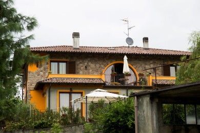 Casa rustica a due piani con pareti arancioni, tetto in terracotta, balcone e giardino sotto un cielo nuvoloso.