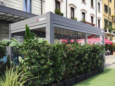 Terrazza esterna di un ristorante con tettoia grigia, siepe verde lussureggiante e ombrelloni rossi su una strada cittadina.