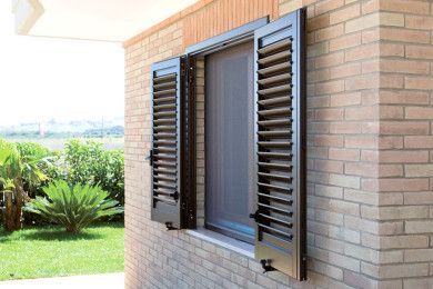 Finestra di una casa in mattoni con persiane marrone scuro, che si apre su un cortile verde brillante.