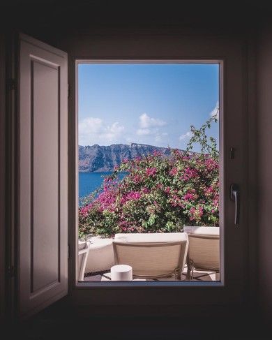 Si apre una porta su un balcone con fiori rosa, mare blu e scogliere in lontananza sotto un cielo luminoso.