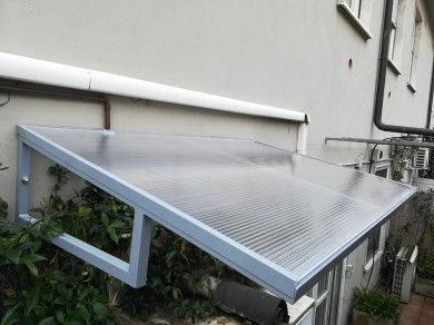 Tenda da sole in metallo con pannelli di copertura traslucidi fissata alla parete dell'edificio, che protegge le piante sottostanti.