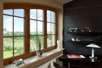Moderno ufficio domestico con ampia finestra ad arco, scrivania scura, mensole e vista sui campi verdi all'esterno.