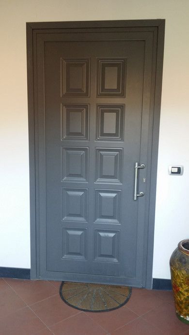 Porta d'ingresso grigia a pannelli con maniglia in metallo, zerbino e piccolo interruttore a parete accanto.