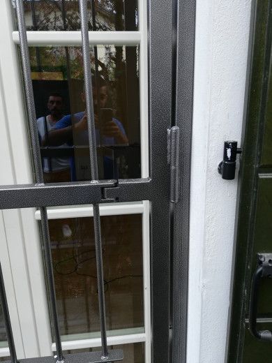 Cancello di sicurezza in metallo davanti a una finestra, con una persona che si riflette debolmente nel vetro.