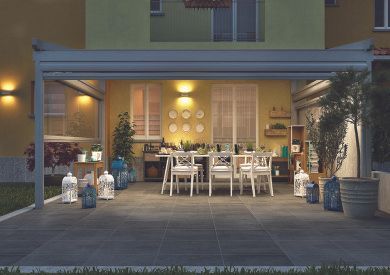 Zona pranzo all'aperto con tavolo e sedie bianchi sotto una terrazza coperta, illuminata da luci calde e lanterne al crepuscolo.