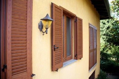 Esterno di una casa gialla con persiane in legno marrone e lanterna a muro accanto a una finestra
