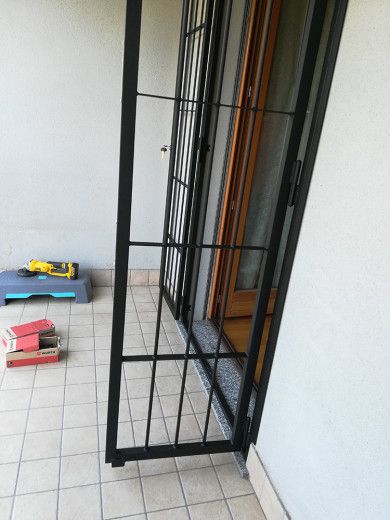 Cancello di sicurezza in metallo nero su un balcone piastrellato, accanto a una parete bianca e porte in legno.