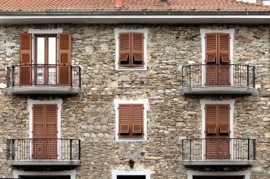 Facciata in pietra dell'edificio con persiane marroni e balconi in ferro battuto sotto un tetto di tegole rosse.