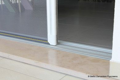 Soglia della porta scorrevole in vetro con binario metallico basso e pavimento piastrellato beige.