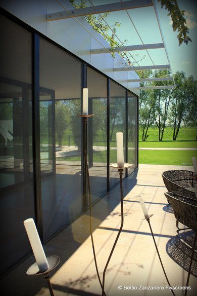 Patio moderno con porte a vetri, pergola bianca, sedie in vimini e vista su un prato verde e alberi.