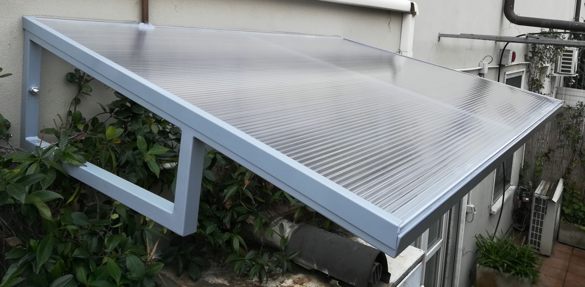 Tenda da sole in metallo con struttura blu sopra un patio esterno, con pannelli di copertura traslucidi ondulati.