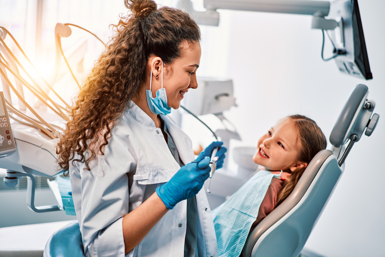 Dentist with curly hair, in blue gloves and mask, smiles at a young patient sitting in a dental chair.