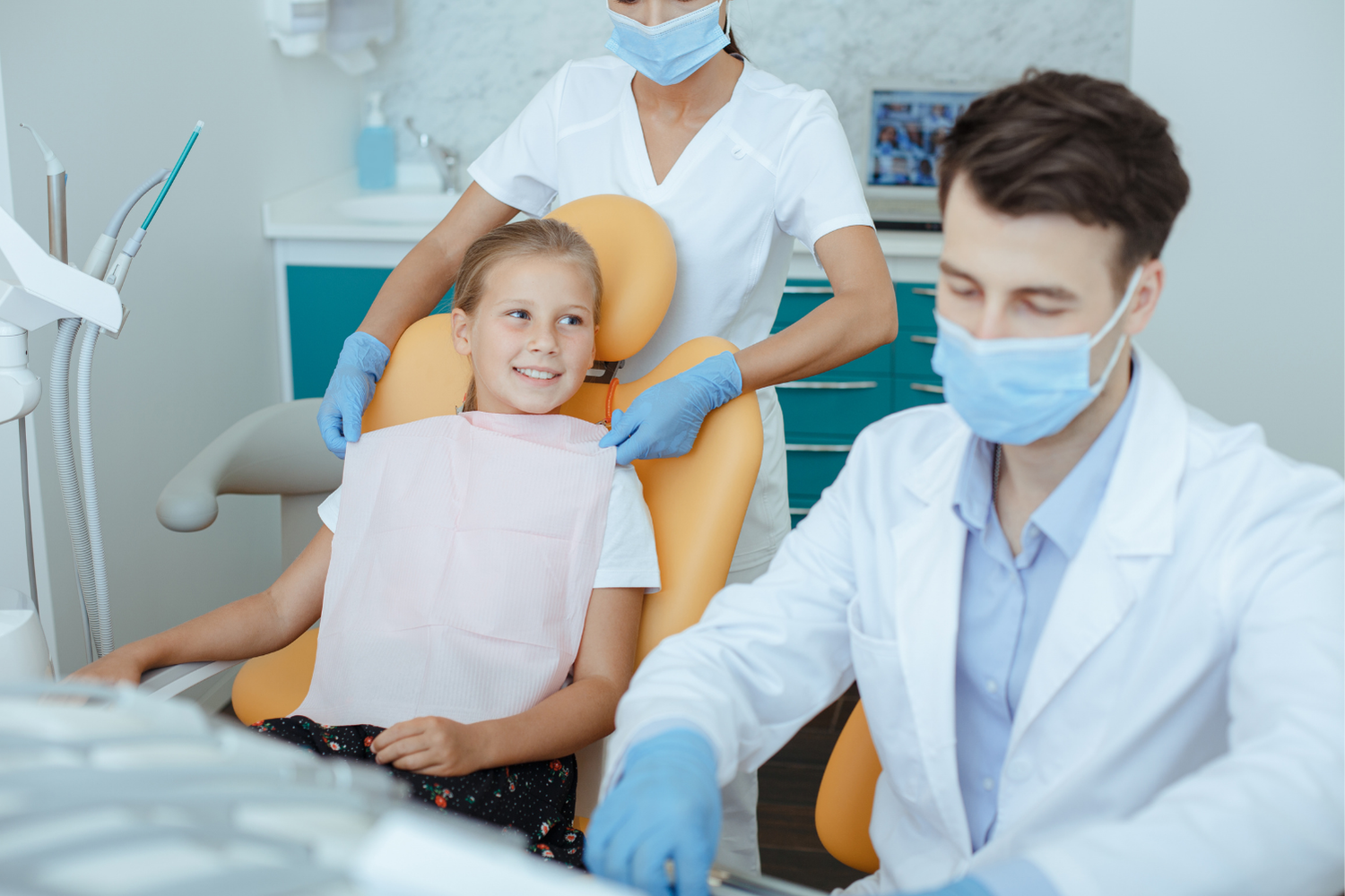 Girl in dental chair, getting a bib put on by a dental assistant, dentist in foreground.