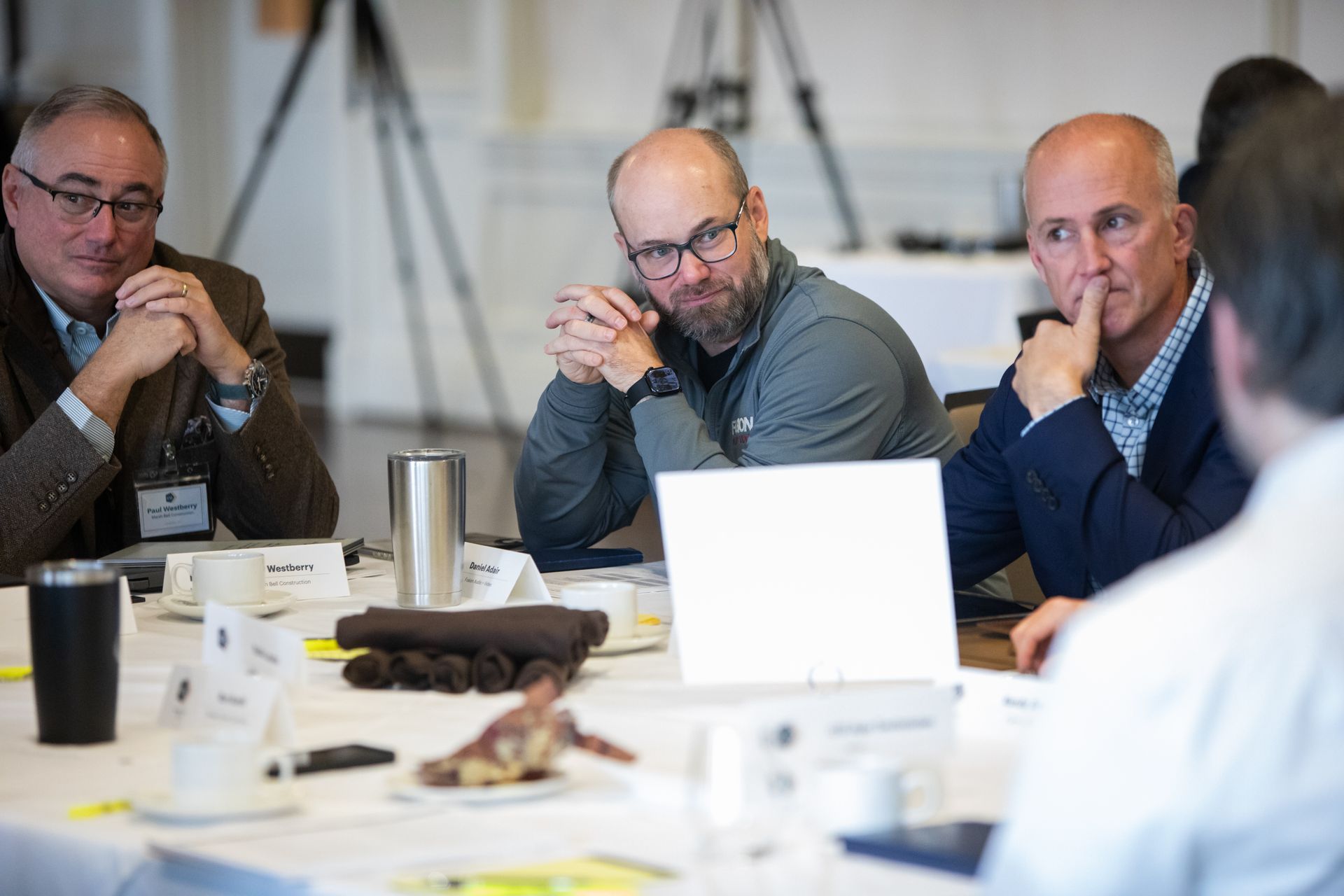 Three people sit at a conference table, looking toward a speaker, with name tags, drinks, and documents on the table.