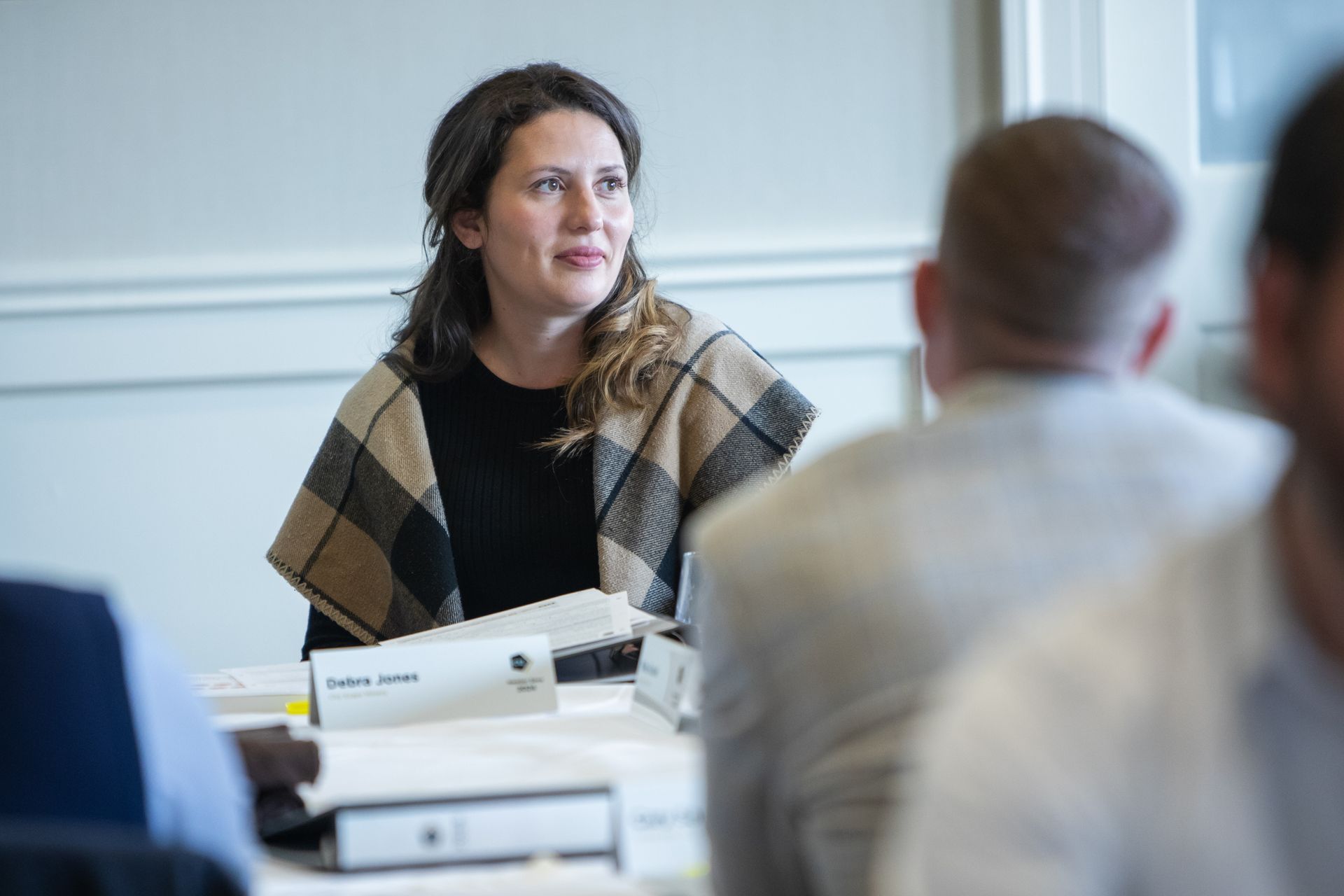 A person in a plaid shawl sits at a meeting table, looking toward the side, with others in the foreground.